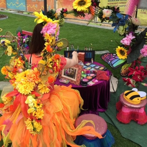 Yello Fairy Face Painting square A girl dressed as a fairy sits at a table with flowers.