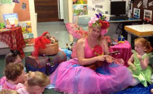 A woman in a vivid pink and purple fairy costume entertains young children in a colorfully decorated classroom.
