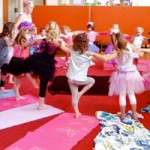 Children in a dance class holding hands and practicing ballet moves in a colorful room with mats on the floor.