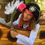A person dressed as a pirate, smiling and holding a curved sword, standing in front of a wooden structure with feathery decorations.